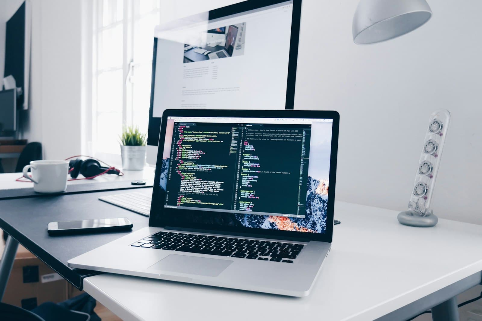 Laptop with code on a white desk with a monitor behind